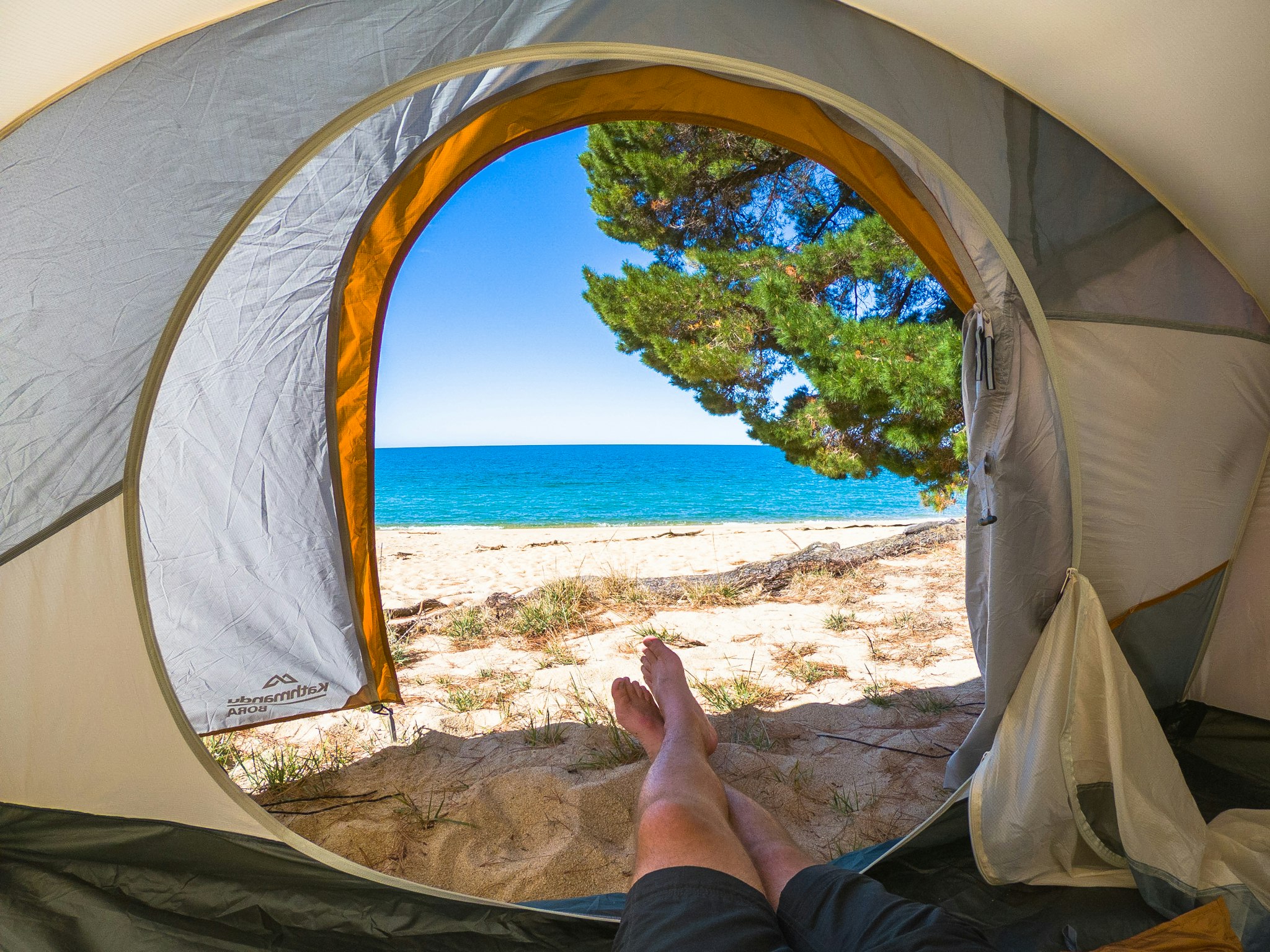 Wake up to views like this one. Image of a pair of legs hanging out of an open tent on a beach. A brilliant blue ocean is in the distance.