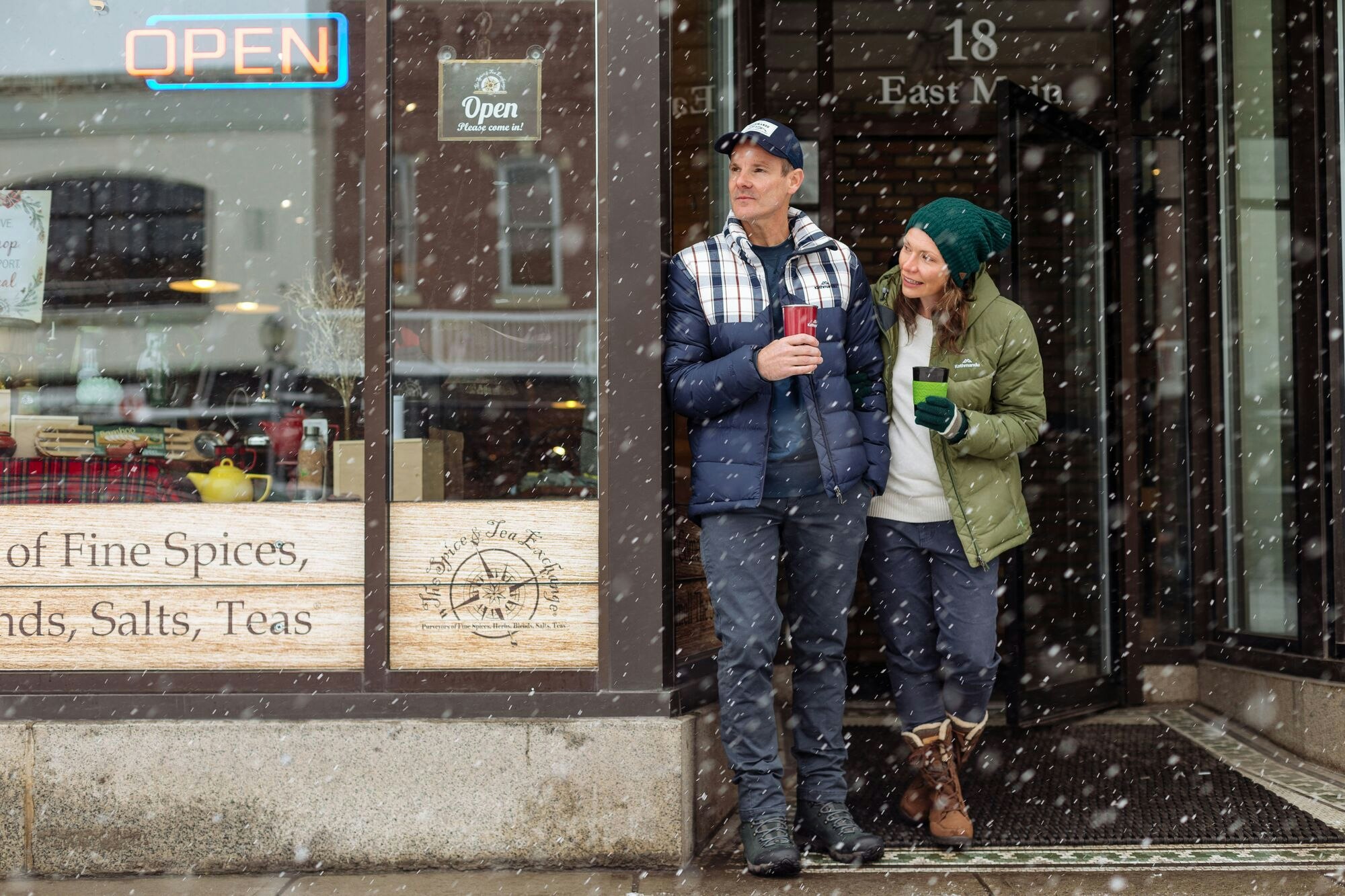 Image of a man and a woman standing in the doorway of a cafe, drinking coffee and watching the snow.